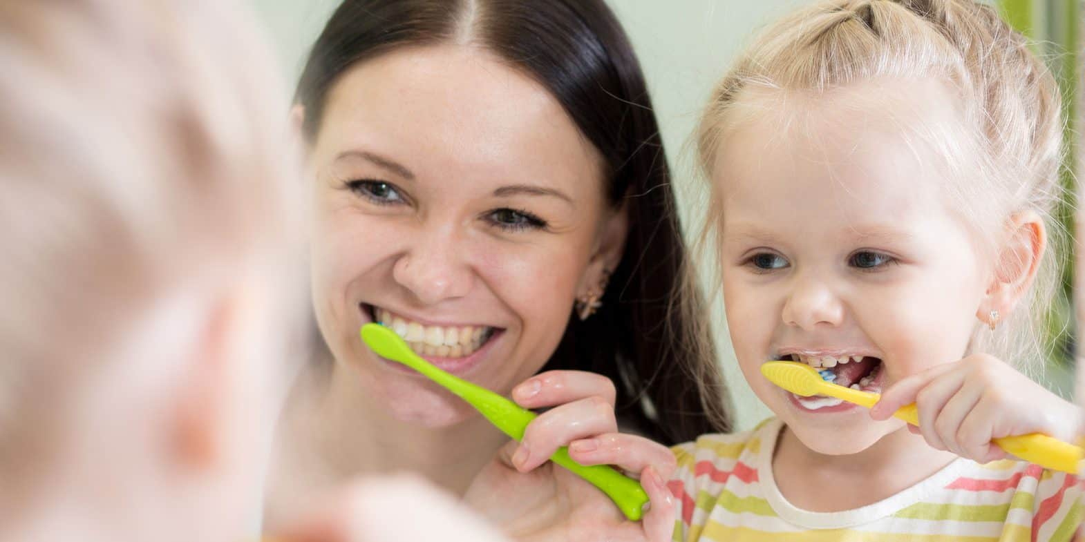 dental-care-tips-main-image A woman and a young girl stand in front of a mirror, smiling and brushing their teeth with green and yellow toothbrushes.