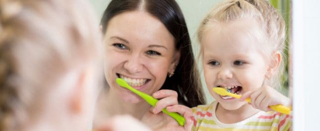 dental-care-tips-main-image A woman and a young girl stand in front of a mirror, smiling and brushing their teeth with green and yellow toothbrushes.