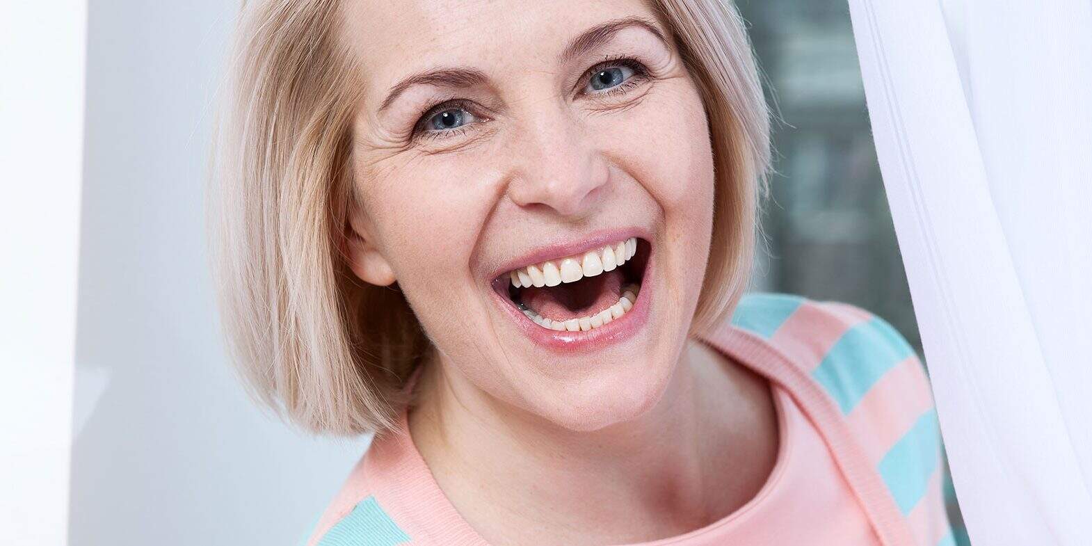 Woman with short blond hair smiling widely while standing near a white curtain, wearing a light pink and blue striped sweater.