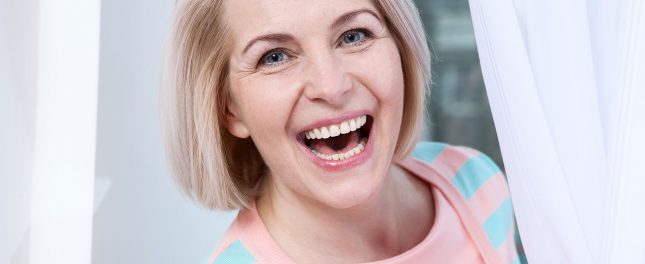 a-healthier-mouth-a-healthier-heart Woman with short blond hair smiling widely while standing near a white curtain, wearing a light pink and blue striped sweater.