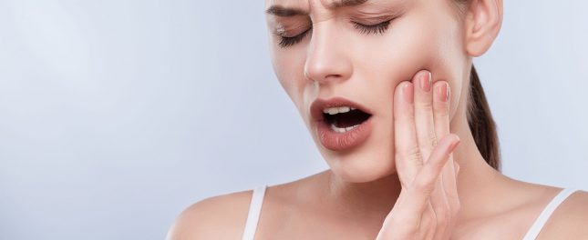 A woman with her mouth slightly open touches her cheek and appears to be in pain, possibly indicating a toothache or jaw discomfort.