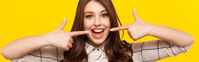 A smiling woman with long brown hair points to her teeth with both hands against a solid yellow background.