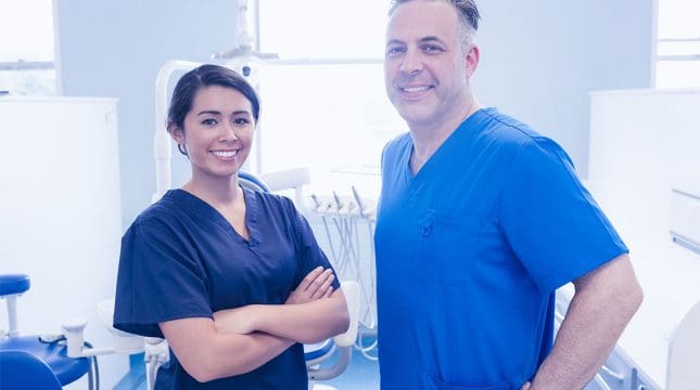 Two dental professionals wearing scrubs stand and smile in a dental clinic, with dental equipment visible in the background.