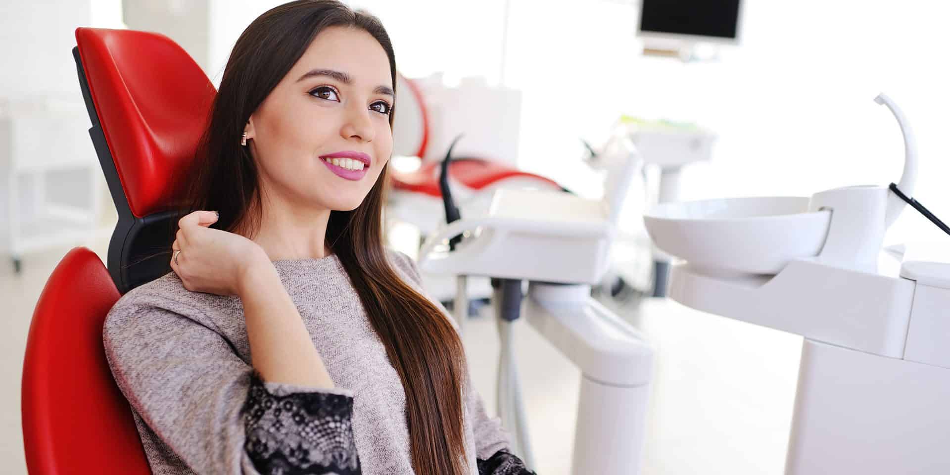 A woman with long brown hair sits and smiles in a red dental chair in a bright dental clinic.