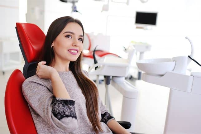 A woman with long brown hair sits and smiles in a red dental chair in a bright dental clinic.
