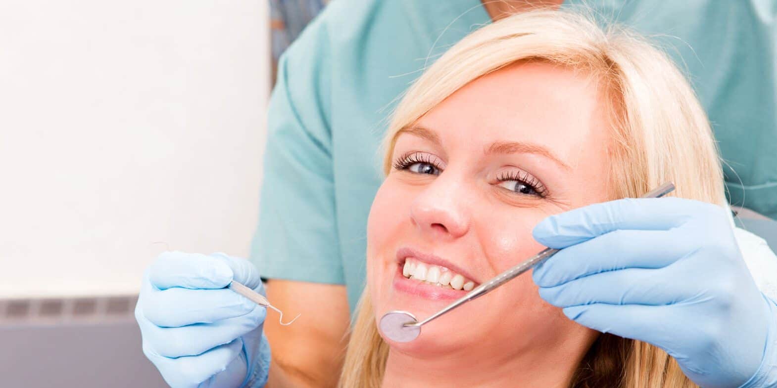 A dentist in blue gloves examines a smiling woman’s teeth using a dental mirror and probe during a dental checkup.