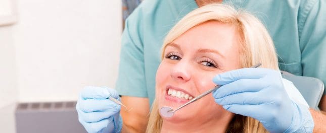 A dentist in blue gloves examines a smiling woman’s teeth using a dental mirror and probe during a dental checkup.