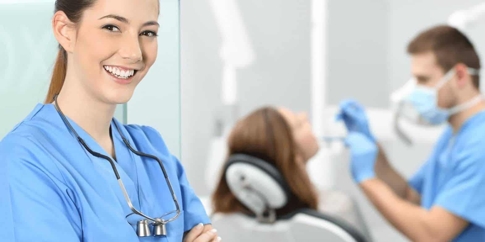 A smiling dental professional stands in the foreground, while another dentist treats a patient in the background of a dental clinic.