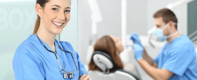 A smiling dental professional stands in the foreground, while another dentist treats a patient in the background of a dental clinic.