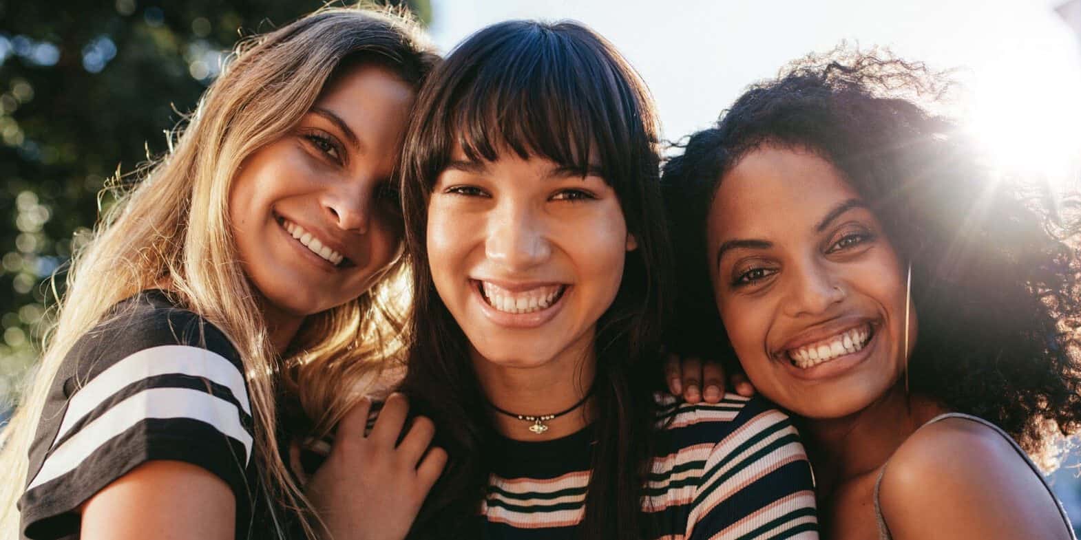 Three young women standing close together outdoors, smiling at the camera with sunlight shining behind them.