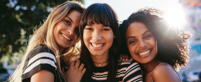 Three young women standing close together outdoors, smiling at the camera with sunlight shining behind them.