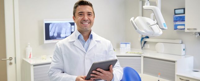 A dentist in a white coat stands in a dental office, holding a tablet and smiling, with dental equipment and an X-ray on a monitor in the background.