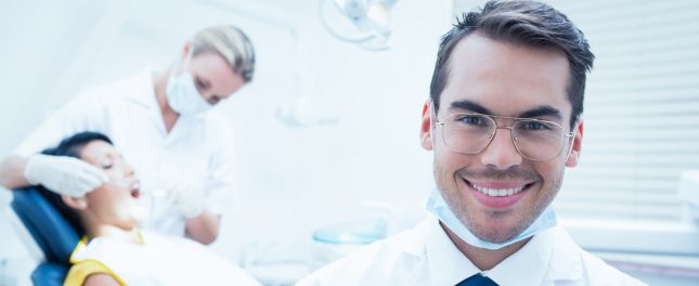 A male dentist wearing glasses and a mask smiles at the camera, while a dental assistant examines a patient in the background.