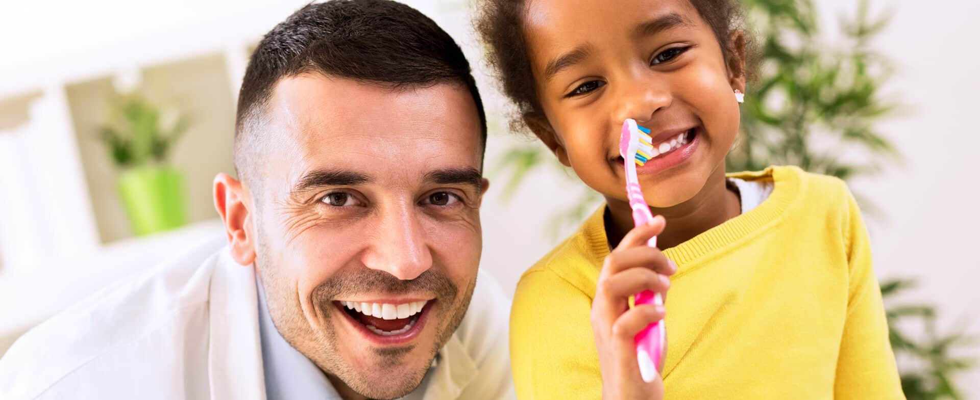A smiling adult and a young child holding a toothbrush pose together indoors, with the child holding the toothbrush near their nose.