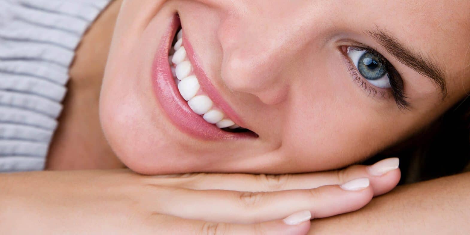 Close-up of a smiling person with blue eyes, resting their head on their hands and looking at the camera.