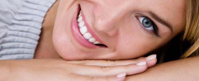 Close-up of a smiling person with blue eyes, resting their head on their hands and looking at the camera.