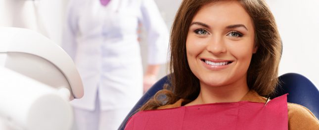 A woman with brown hair sits in a dental chair, wearing a protective bib, and smiles at the camera. Experience comfort and ease with Sedation Dentistry Georgia while a dentist stands blurred in the background.