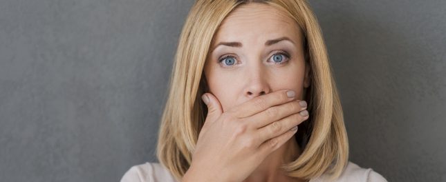A woman with blonde hair looks surprised and covers her mouth with her hand, standing in front of a gray background.