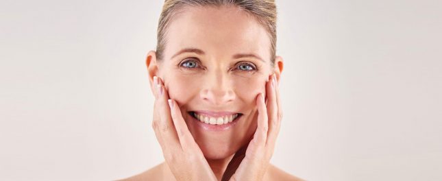 A middle-aged woman with blond hair is smiling and gently touching both sides of her face with her hands against a plain, light background.