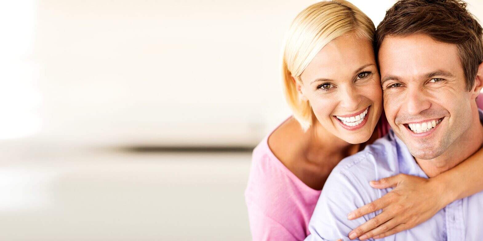 Smiling woman hugs a smiling man from behind, both looking at the camera in a brightly lit indoor setting.