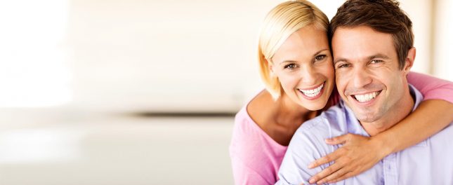 Top-3-Benefits-Of-Professional-Teeth-Whitening-Treatment Smiling woman hugs a smiling man from behind, both looking at the camera in a brightly lit indoor setting.