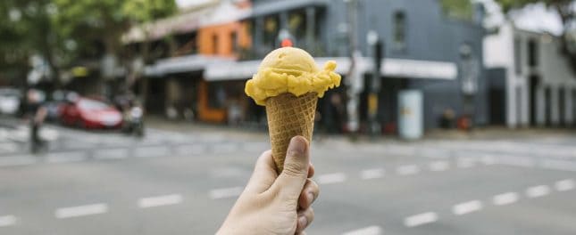 A hand holds an ice cream cone with yellow ice cream and a red topping, with a city street and buildings in the blurred background.