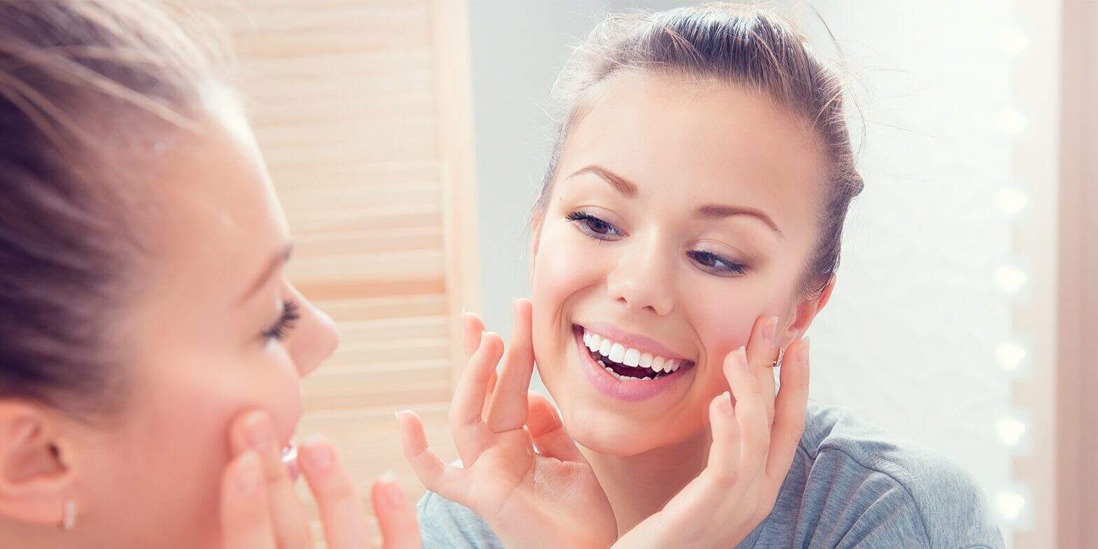 relief-for-sensitive-teeth Young woman smiling and touching her cheeks while looking at herself in the bathroom mirror.