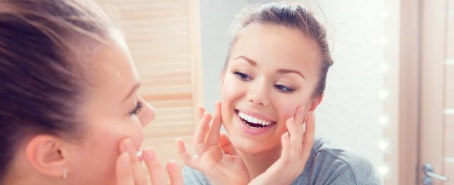 relief-for-sensitive-teeth Young woman smiling and touching her cheeks while looking at herself in the bathroom mirror.