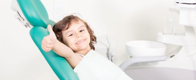 A young child sits in a dental chair, smiling and giving a thumbs-up, with dental equipment visible in the background.