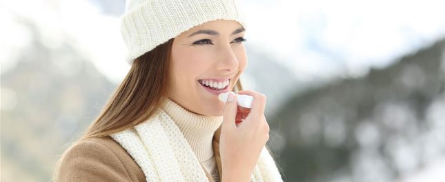 A woman wearing a knit hat and scarf applies lip balm outdoors in a snowy, mountainous setting.
