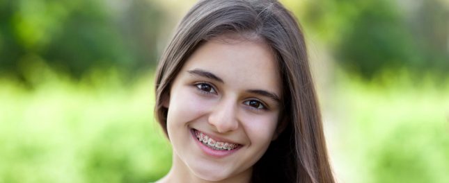 A young person with long brown hair and braces smiles at the camera with a blurred green outdoor background.