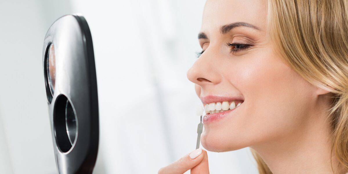 A woman smiling at herself in a handheld mirror while brushing her teeth with a toothbrush.