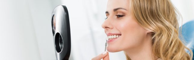 A woman smiling at herself in a handheld mirror while brushing her teeth with a toothbrush.