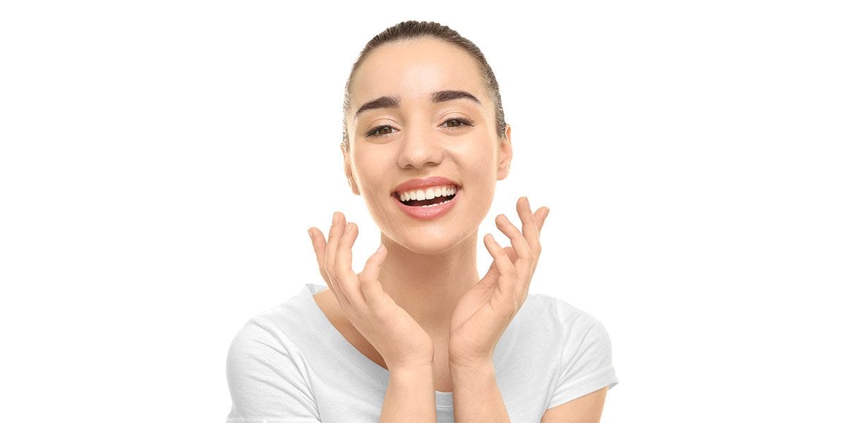 Young woman with tied-back hair smiling and touching her face with both hands, wearing a white shirt against a white background.