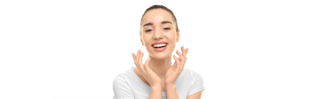 Young woman with tied-back hair smiling and touching her face with both hands, wearing a white shirt against a white background.