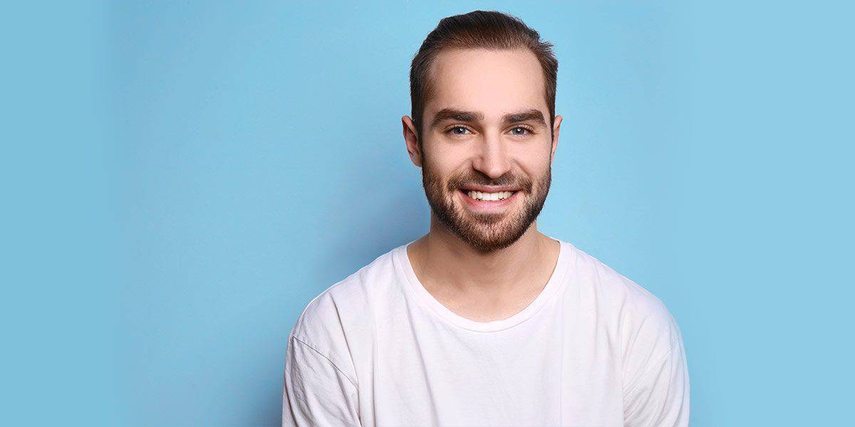 A man with short brown hair and a beard smiles at the camera while wearing a white t-shirt, standing against a plain blue background.