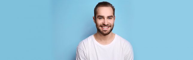 A man with short brown hair and a beard smiles at the camera while wearing a white t-shirt, standing against a plain blue background.