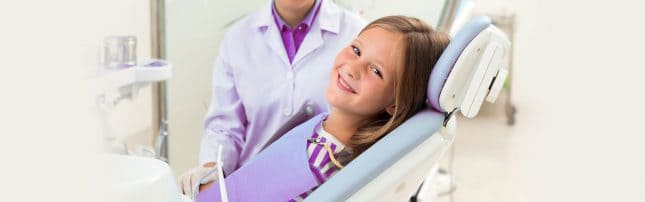 A young girl sits in a dental chair, smiling at the camera, while a dental professional in a lab coat stands beside her.