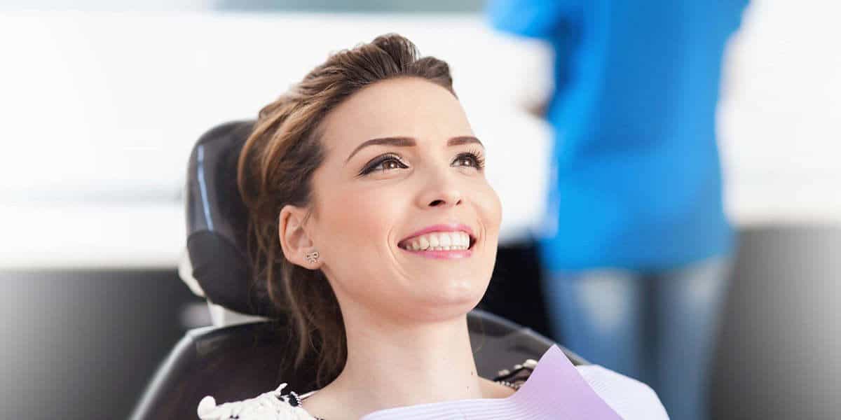 A woman sits in a dental chair smiling, wearing a dental bib, with a dental professional standing in the background.