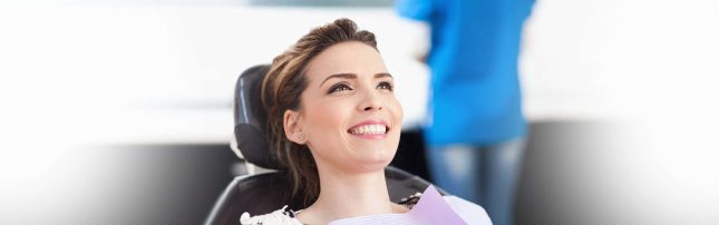 A woman sits in a dental chair smiling, wearing a dental bib, with a dental professional standing in the background.