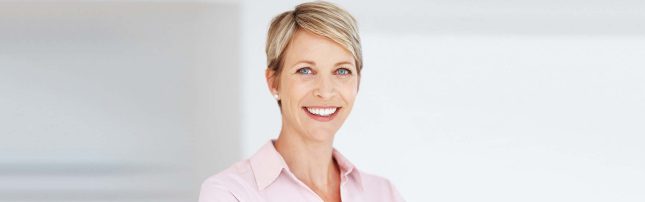 A woman with short blonde hair and blue eyes is smiling, wearing a light pink collared shirt, standing in a bright, plain background.