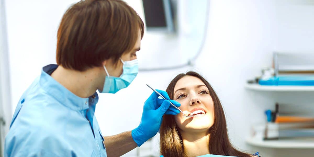 A dentist wearing a mask and gloves examines a woman’s teeth in a brightly lit dental clinic.