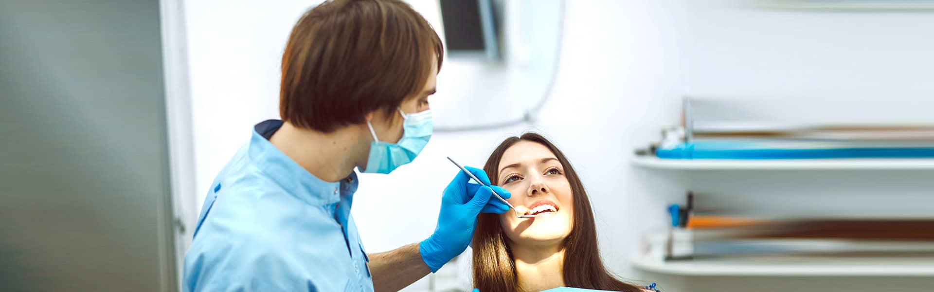 A dentist wearing a mask and gloves examines a woman’s teeth in a brightly lit dental clinic.