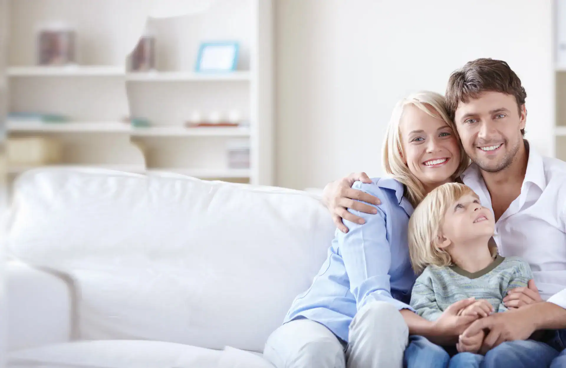 A man, woman, and young boy sit closely together on a white couch in a living room, smiling at the camera.