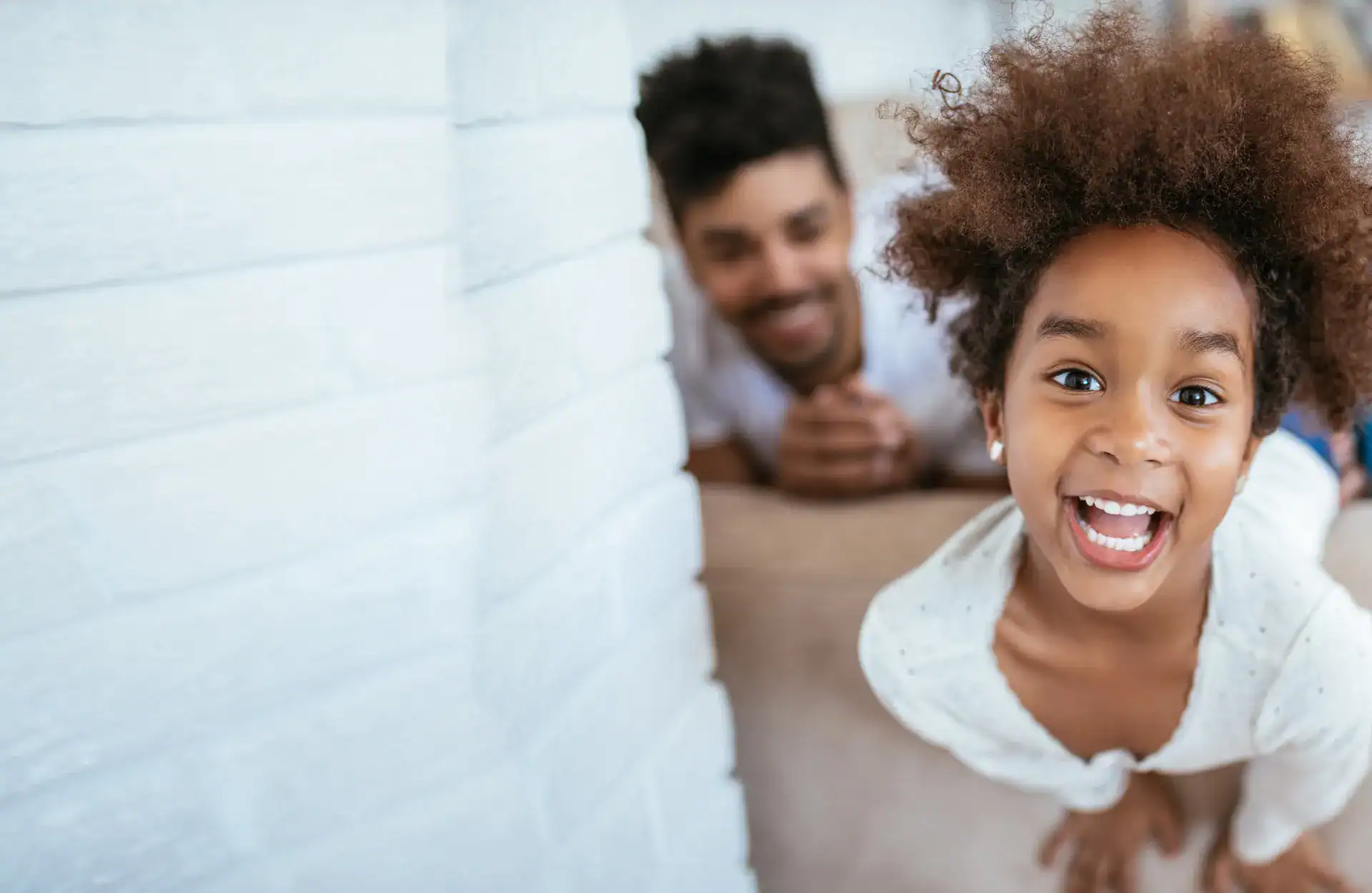 A smiling child leans toward the camera while an adult, slightly blurred, watches from behind in a bright indoor setting.