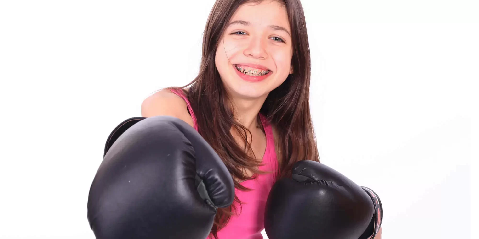 Smiling girl with long brown hair and braces, thanks to Complete Family Dental Services East Point GA, wears a pink tank top and black boxing gloves, posing with fists raised against a white background.