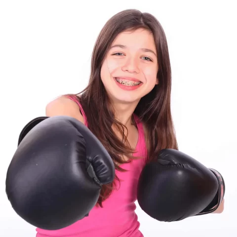 Smiling girl with long brown hair and braces, thanks to Complete Family Dental Services East Point GA, wears a pink tank top and black boxing gloves, posing with fists raised against a white background.