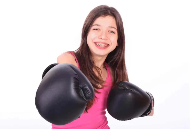 Smiling girl with long brown hair and braces, thanks to Complete Family Dental Services East Point GA, wears a pink tank top and black boxing gloves, posing with fists raised against a white background.